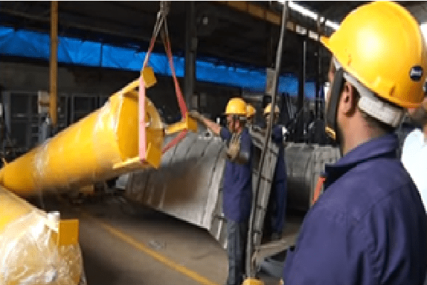 Factory workers in yellow hard hats guide a large yellow turbine blade with straps in a factory setting.