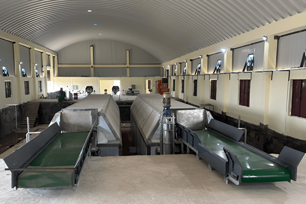 Industrial processing facility with two green conveyor belts feeding into a central processing line under a high arched ceiling in a long warehouse.